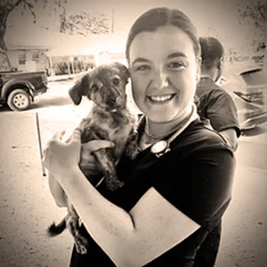 Veterinarian Dr. Karli Collins smiles while holding a small puppy outside near parked cars.