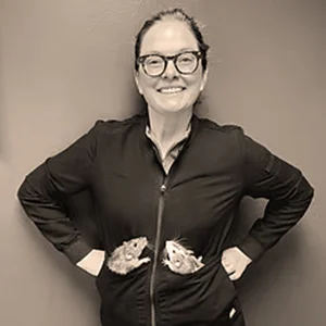Veterinarian Dr. Samantha Coe smiles with hands in her jacket pockets, which feature images of two small animals.
